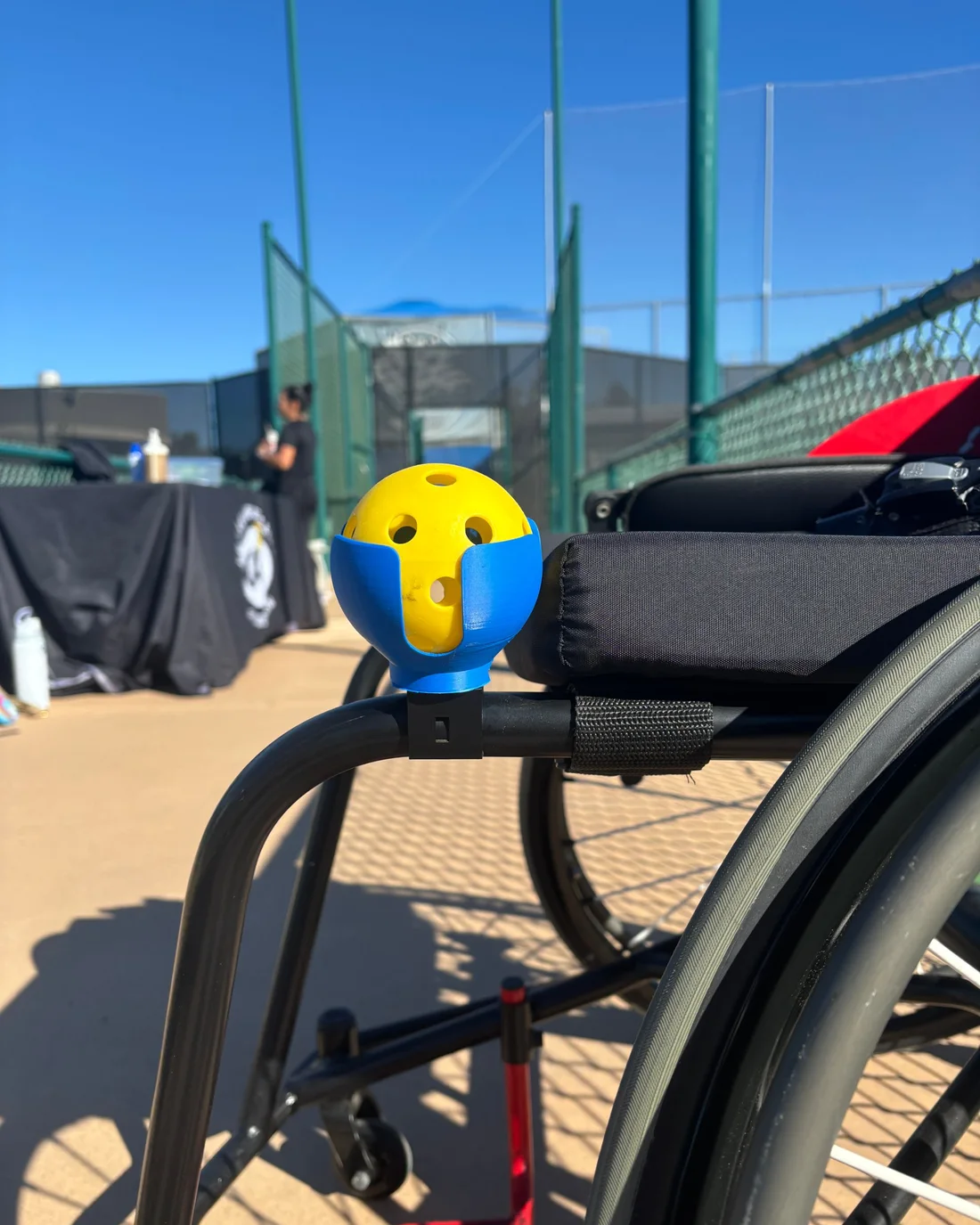 A blue PickleDock holding a yellow pickleball, clamped to the frame of a sports wheelchair next to the court