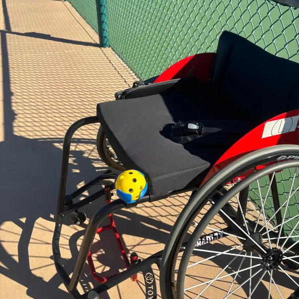 A sports wheelchair by the court fence with PickleDock mounted near the front of the seat